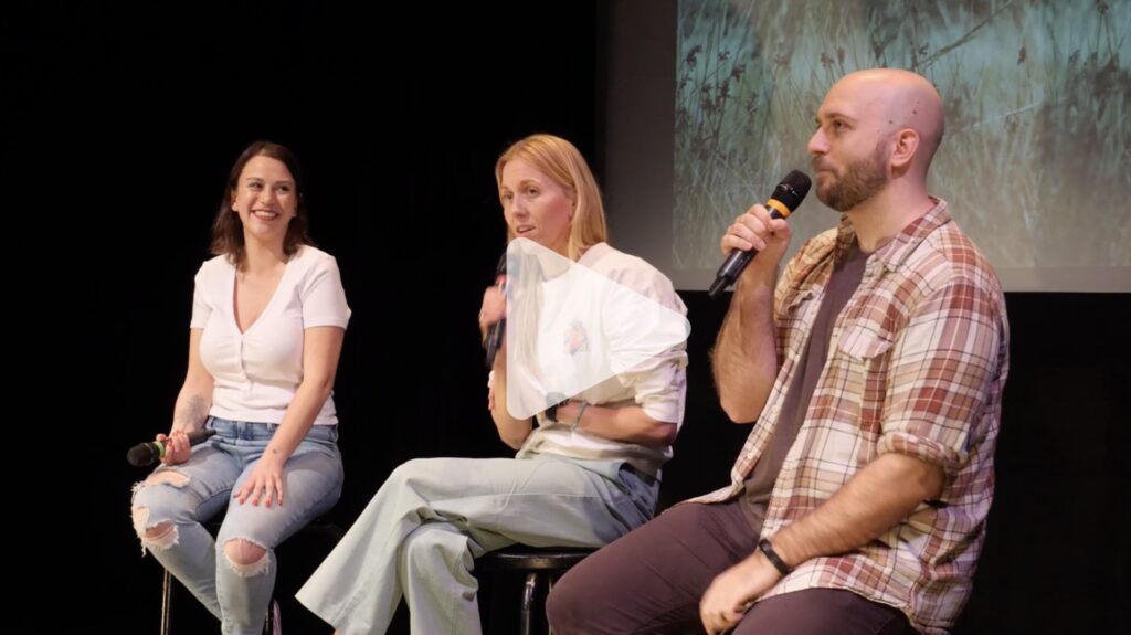 Two women and a man sit on stools in front of a film screen having a conversation while holding microphones