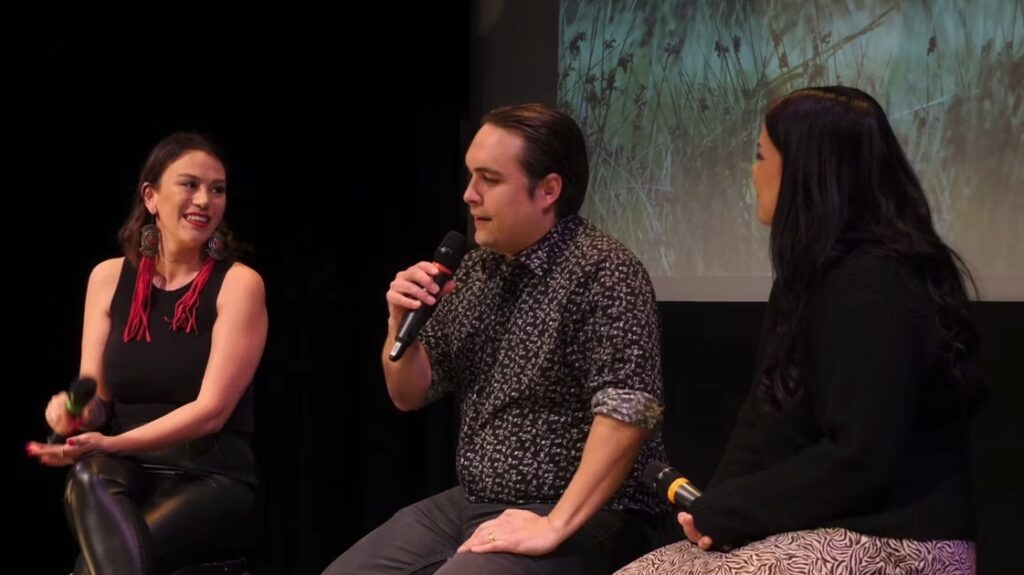 Two women and a man sit on stools in front of a film screen having a conversation while holding microphones