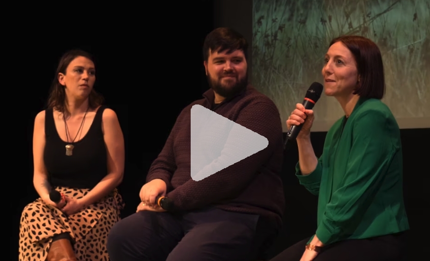 A woman in black tank top, a bearded man in a long sleeve dress shirt and a woman in a green shirt sit in front of a film screen