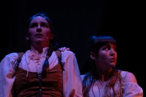 Two women in shakespearian costumes in shadowy pink and purple light with worried expressions.