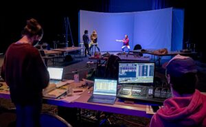 technicians at a table with laptops in the foreground with rehearsal for I Am The Cheese on stage in the distance