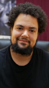 Jackson, a mixed-race African Nova Scotian man, with a tan complexion, curly hair and beard, looks into the camera with a friendly, warm smile.