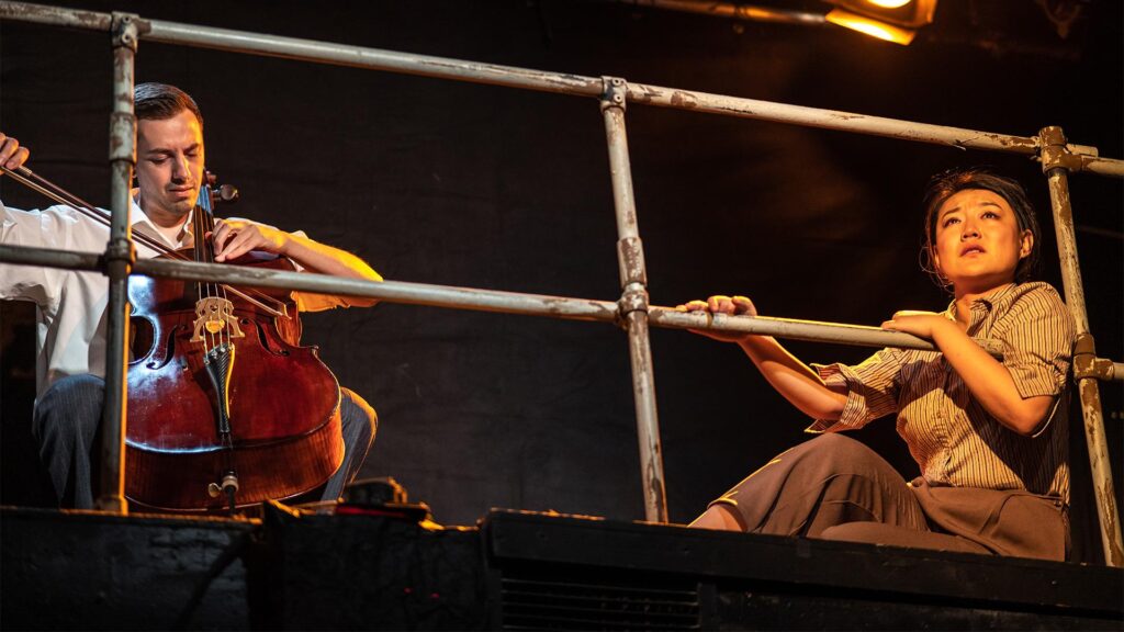 A man plays the cello beside a woman seated on the ground with a wistful look on her face. They are behind a metal railing with large openings and lit with warm yellow light from above.