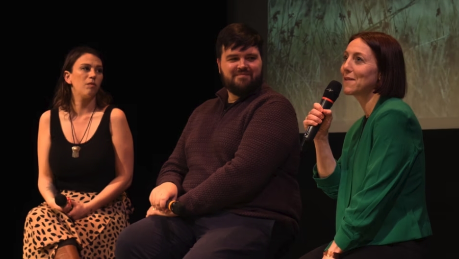 A woman in black tank top, a bearded man in a long sleeve dress shirt and a woman in a green shirt sit in front of a film screen