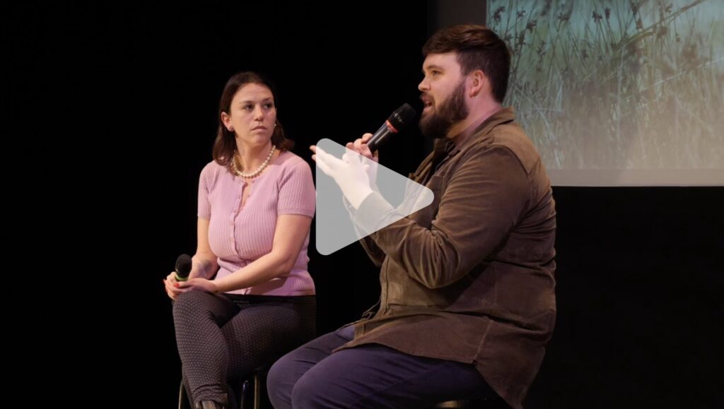 A woman with shoulder length brown hair and a pink sweater and a bearded man with short brown hair and a brown long sleeve shirt sit on stools holding microphones in front of a film screen.